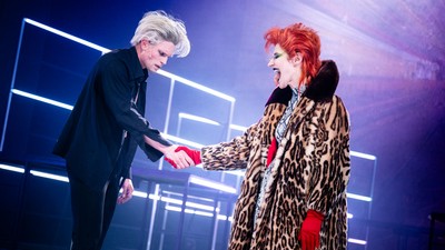 Zwei Darsteller auf der Bühne, die sich die Hände schütteln – einer mit platinblondem Haar in schwarzer Kleidung, der andere mit rotem Haar in einem Leopardenmantel.