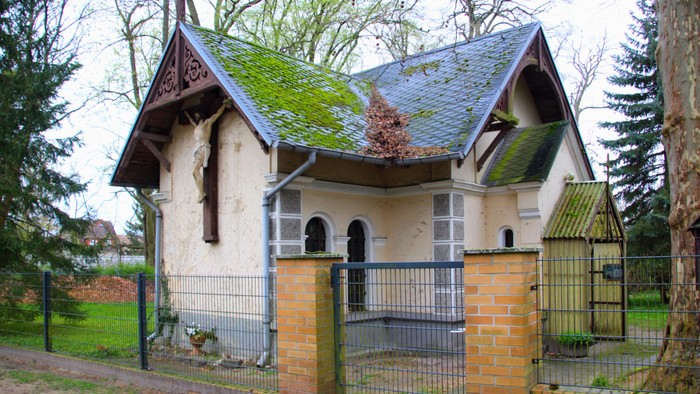 Kleines Haus mit steilem Dach und Moos, umgeben von einem Metallzaun.