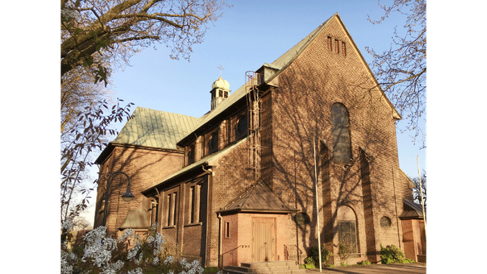 Große Steinkirche mit hohen Fenstern und einem grünen Dach, umgeben von Bäumen und Sträuchern.