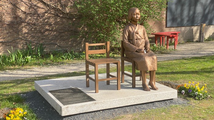 Bronzestatue einer auf einer Holzbank sitzenden Frau in einem Garten.