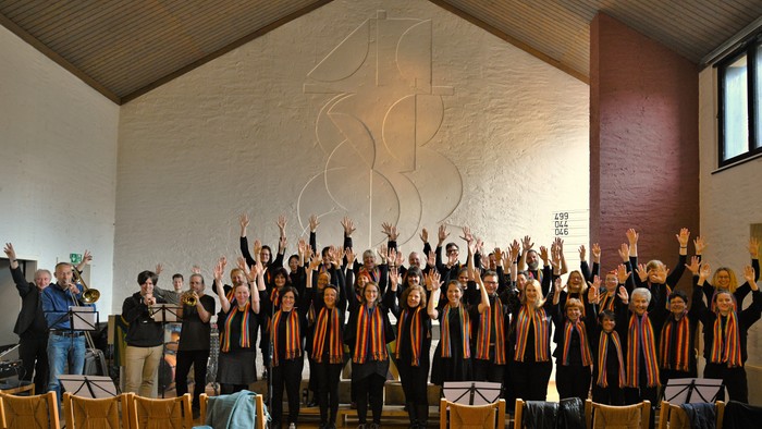 Ein großer Chor singt in einer Kirche, alle Mitglieder tragen orange-schwarze Outfits und heben die Arme, während ein Mann Notenblätter hält.