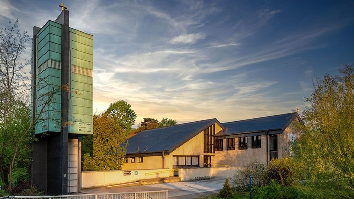 Modernes Gebäude mit grünem Turm und großen Fenstern, umgeben von Bäumen und Büschen unter blauem Himmel
