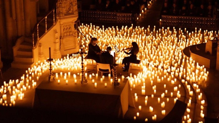 Two people sit among numerous lit candles in a dimly lit area.