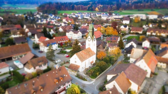 Dorf mit Kirche und umliegenden Häusern, umgeben von Bäumen und Feldern