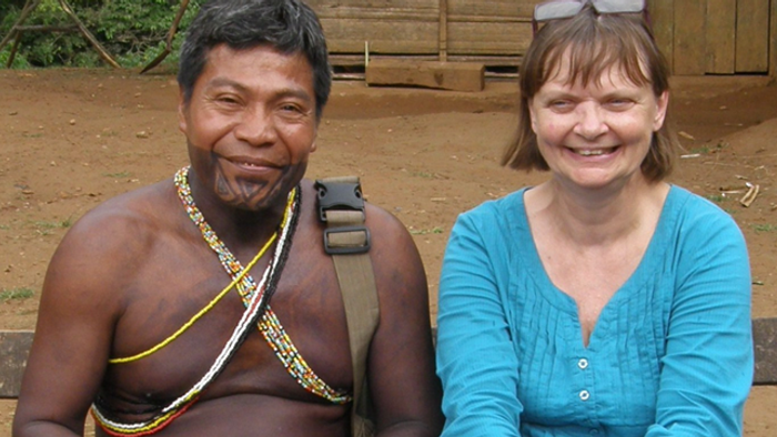 To personer smiler, den ene iført traditionelle oprindelige smykker og den anden i afslappet påklædning, udendørs.