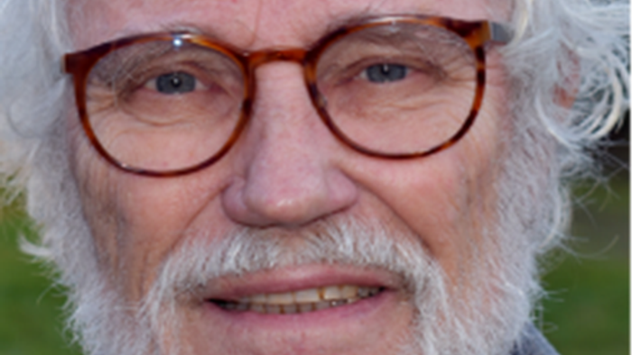 Ældre mand med krøllet hvidt hår, overskæg og briller, der smiler udendørs.