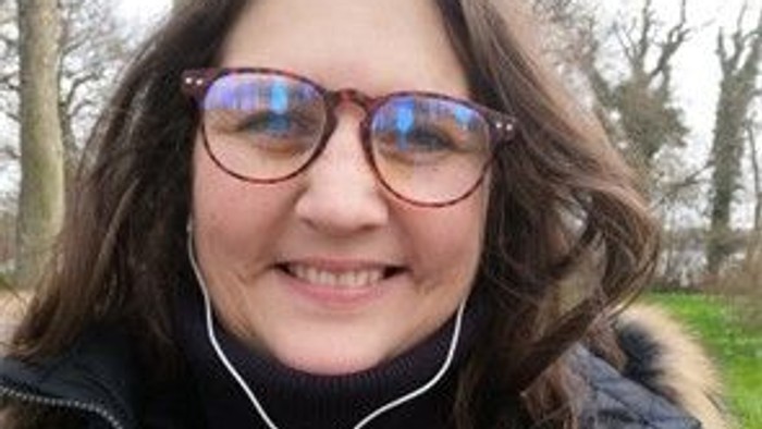 En person med briller, hovedtelefoner og en pelsforet jakke smiler udendørs i en parkagtig omgivelse.