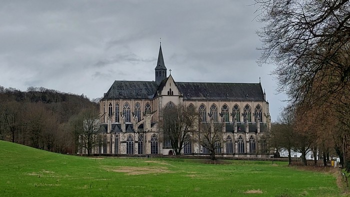 Großes gotisches Gebäude mit spitzem Turm auf grünem Hügel, umgeben von Bäumen unter bewölktem Himmel