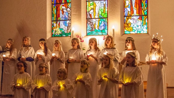 En gruppe børn i hvide klerer med lysestake står foran farverige vinduer i en kirke