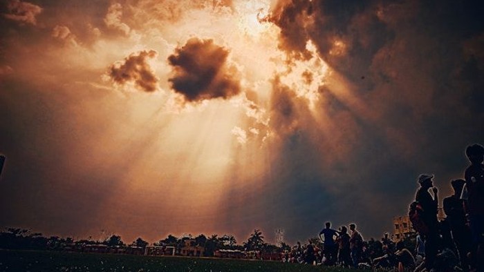 Menschen versammeln sich auf einem Feld unter einem dramatischen Himmel mit Sonnenstrahlen.