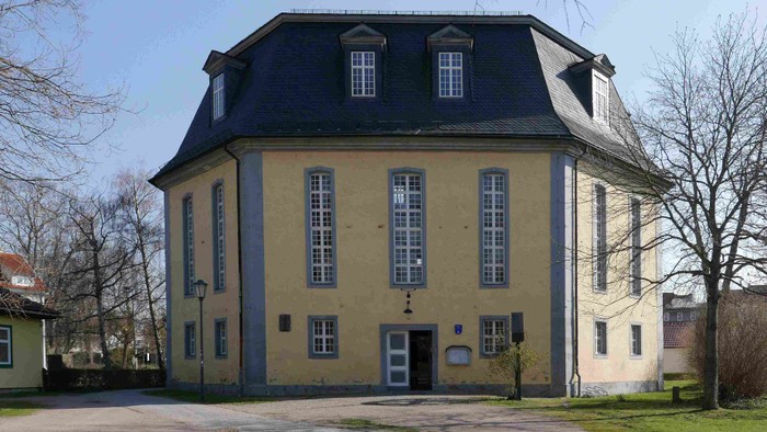 Das Bild zeigt ein großes, gelbes Gebäude mit schwarzem Dach und mehreren Fenstern.