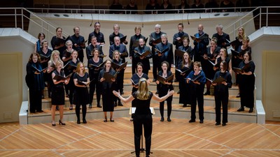 Ein großer Chor tritt gemeinsam in einem prächtigen Konzertsaal auf, während das Publikum von der Empore aus zuschaut.