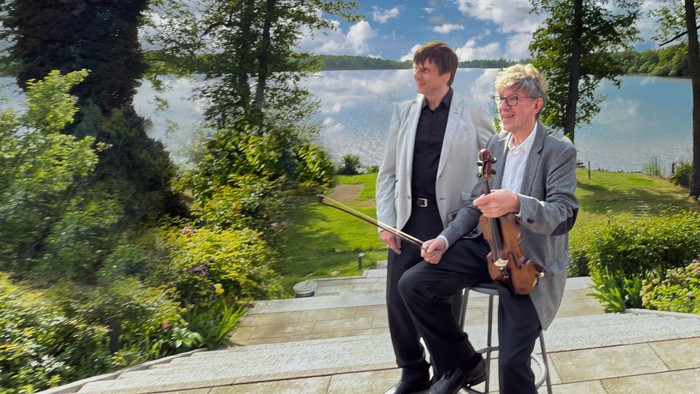 Zwei Musiker, einer mit Notenblättern und der andere mit einer Geige, stehen im Freien auf einer steinernen Terrasse.