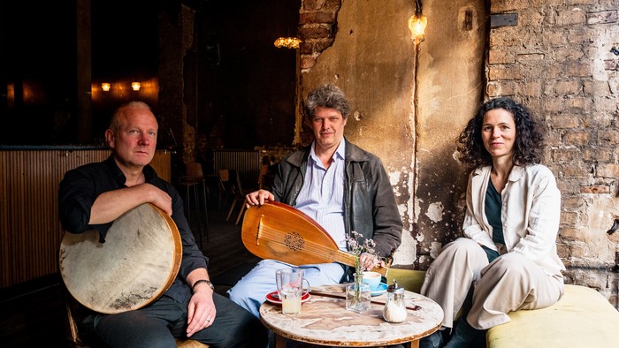 Drei Musiker sitzen gemeinsam an einem rustikalen Tisch mit Instrumenten und genießen eine gemütliche Atmosphäre in einem Innenraum.