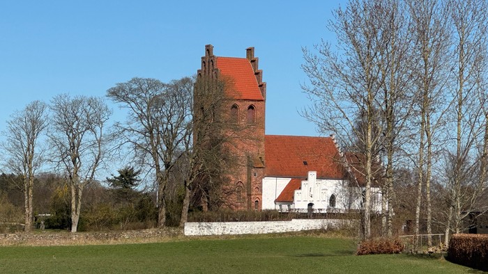 Historisk stenkirke med rødt teglstenstag placeret i et fredeligt, grønt landskab omgivet af nøgne træer.