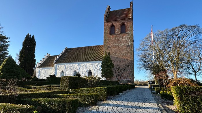 Historisk kirke med hvide mure og murstens tårn, omgivet af veltrimmede hække og en brostensbelagt sti.