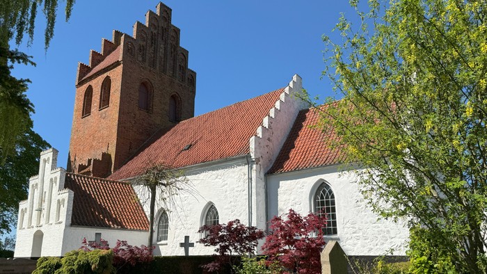 Et malerisk hvid kirke med røde teglstenstag og et murstens tårn, omgivet af frodig grønne omgivelser.