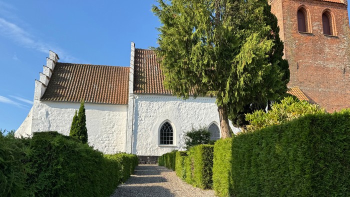 Hyggelig hvid kirke med grussti og veltrimmede hække, omgivet af frodig grønne omgivelser.