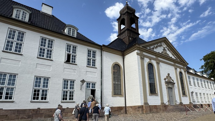 En gruppe går forbi en historisk hvid kirke med et klokketårn og tilstødende bygninger i klassisk stil.