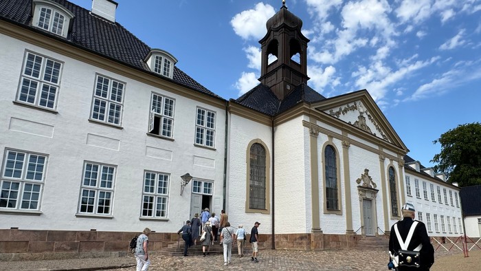 Historisk hvid kirke og tilstødende bygning med mennesker, der går på en brostensbelagt gårdsplads under klar himmel.