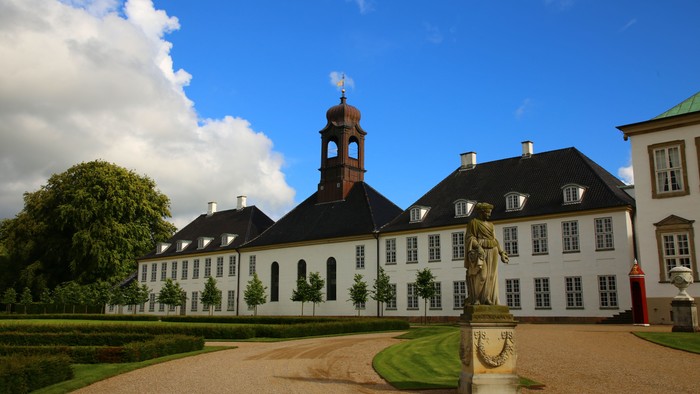 Elegant historisk bygning med symmetrisk arkitektur, central statue og anlagt gårdhave under en klar himmel.