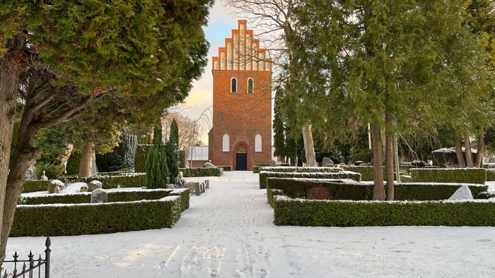 En sneklædt sti fører til et rødt murstårn omgivet af træer og hække.