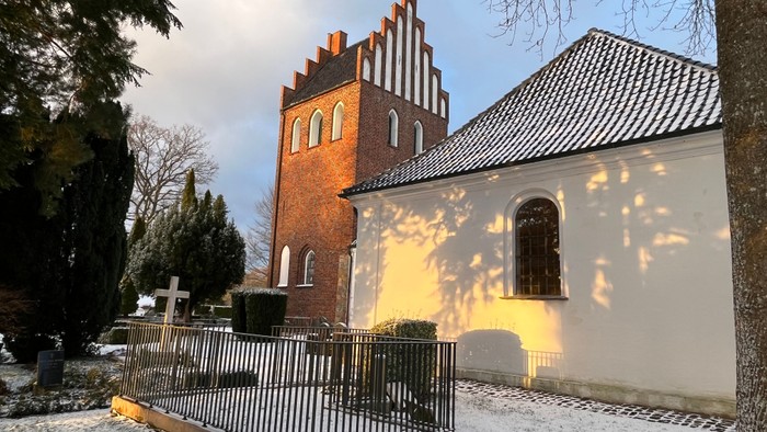 En snedækket kirkegård med en rød murstenskirke og et hvidt sidebygning.