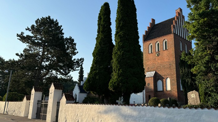 Murstensbygning med høje træer og hvid mur.