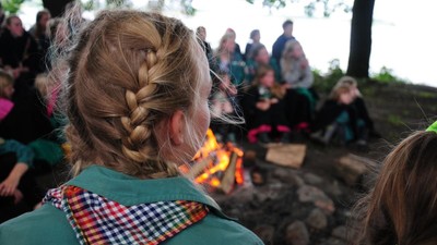 En gruppe børn sidder rundt på en bål, en pige med to frisyrer i baghovedet.