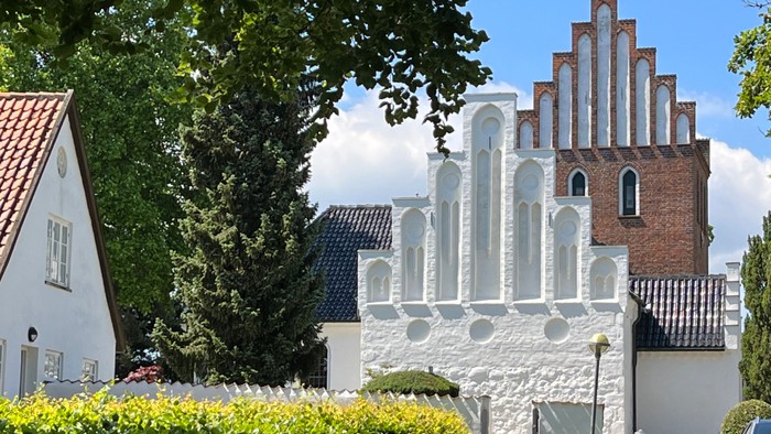 Historisk kirke med murstens tårn og hvid facade omgivet af charmerende, velholdte bygninger og grønne områder.