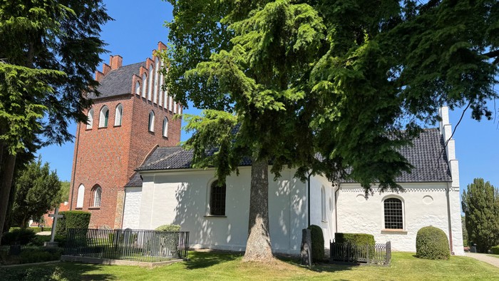 Charmerende hvid kirke med et rødt murstens tårn, omgivet af frodig grønne områder og velplejede græsplæner.
