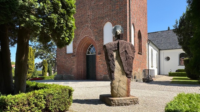 En statue foran en rød murstenskirke med høje træer og en græsplæne