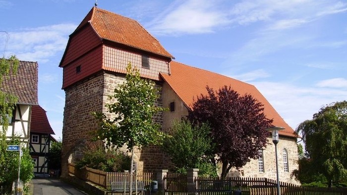 Historisches Gebäude mit rotem Dach und Turm, umgeben von Bäumen und einem Zaun.