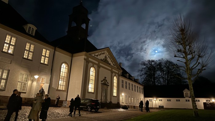 En stor belyset bygning med flere vinduer og en klokketårn, omgivet af en gruppe mennesker og et auto.