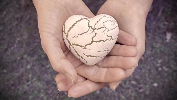 Hände halten sanft einen rissigen herzförmigen Stein, der Widerstandsfähigkeit und Liebe symbolisiert.