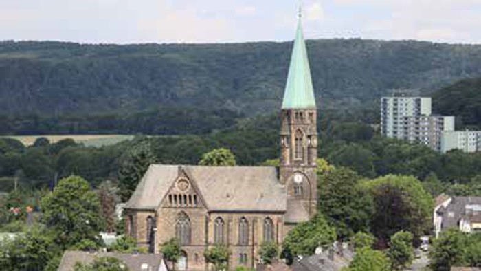 Eine Kirche mit hohem Turm in einem grünen, hügeligen Gebiet mit Wohnhäusern und hohen Gebäuden im Hintergrund.