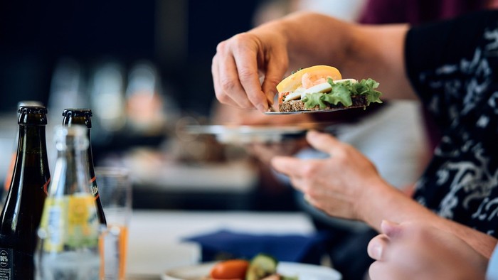 Person, der holder en sandwich, mens de bruger en smartphone ved et spisebord med drikkevarer og mad.