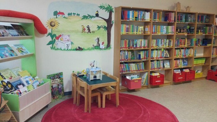 Kinderbibliothek mit bunten Bücherregalen, Tisch und Teppich.