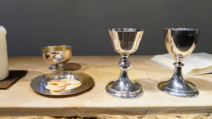 Religiöser Altaraufbau mit Kerze, Brot und zwei zeremoniellen Weinkelchen auf einer Holzoberfläche.