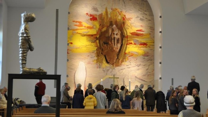 En gruppe mennesker sidder i en kirke, hvor der er en stor skikkelse og et maleri på veggen.