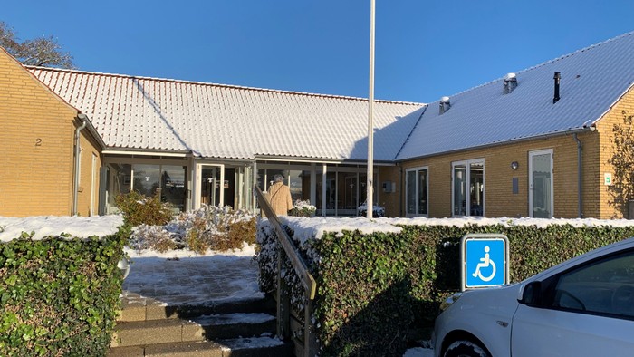 En hvid bil parkeret foran et hus med handicapparkeringstegn.