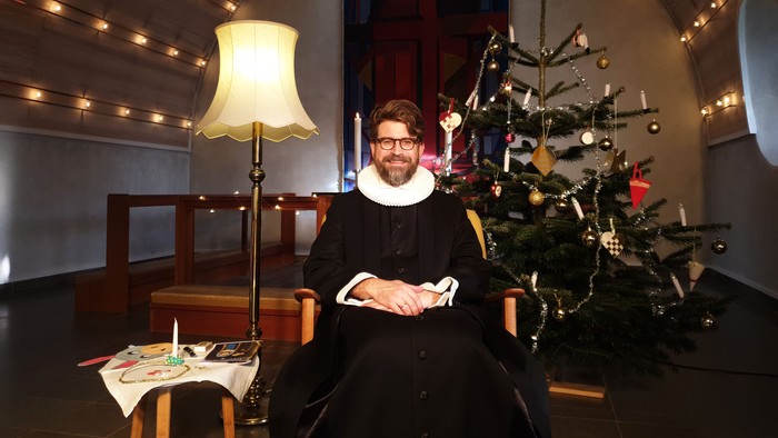 En mand i svart dragt sidder ved juletree og smiler