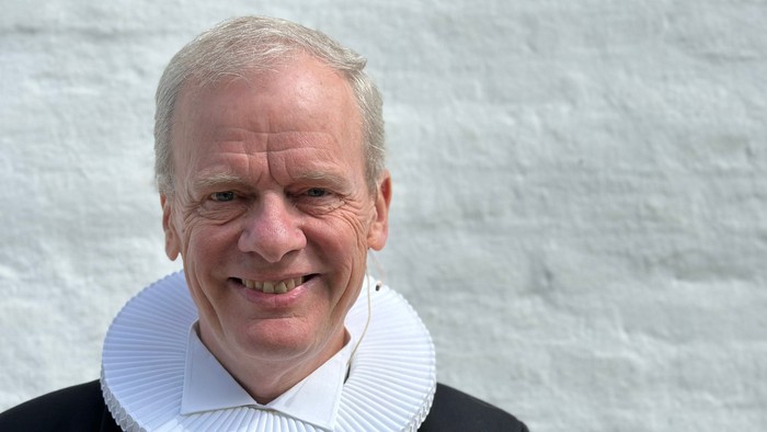 En smilende mand med hvid krage og sort jakke står foran en grå mur.