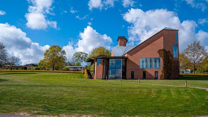 Et moderne hus med træfacader og store vinduer, omgivet af græs og træer under en klar blå himmel.
