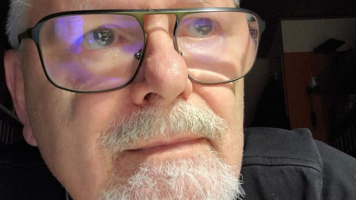 En mand med briller og overskæg iført en sort skjorte, der tager et selfie med et farverigt lysrefleks på sine brilleglas.