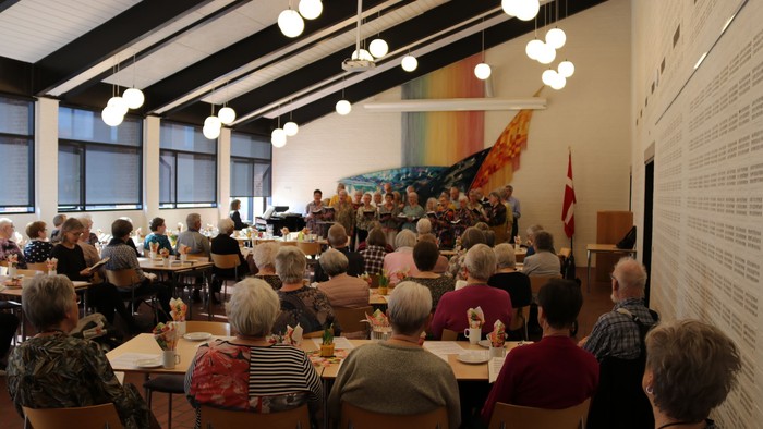 En gruppe mennesker samlet i en rummelig spisesal, hvor de nyder et fælles måltid og samtale.