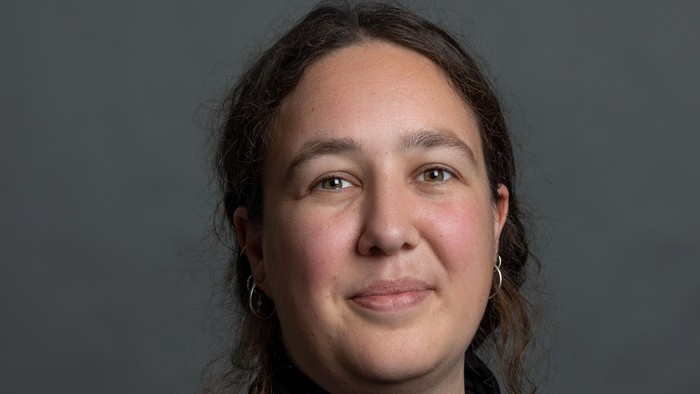 En kvinde med mørkt skulderlangt hår og store øreringe, iført en mørk top, smiler blidt mod en grå baggrund.