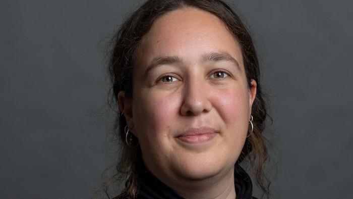 En kvinde med mørkt skulderlangt hår og store øreringe, iført en mørk top, smiler blidt mod en grå baggrund.