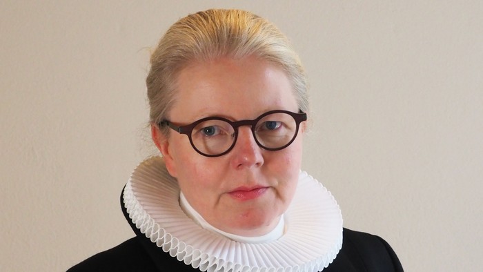 En kvinde med blond hår og briller bærer en sort jakke med hvid krage.