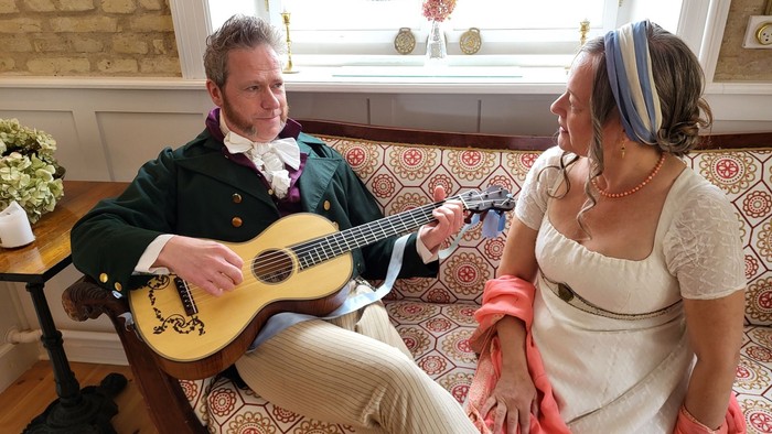 En mand i vintage-tøj spiller guitar, mens en kvinde i en elegant kjole lytter opmærksomt ved siden af ham.